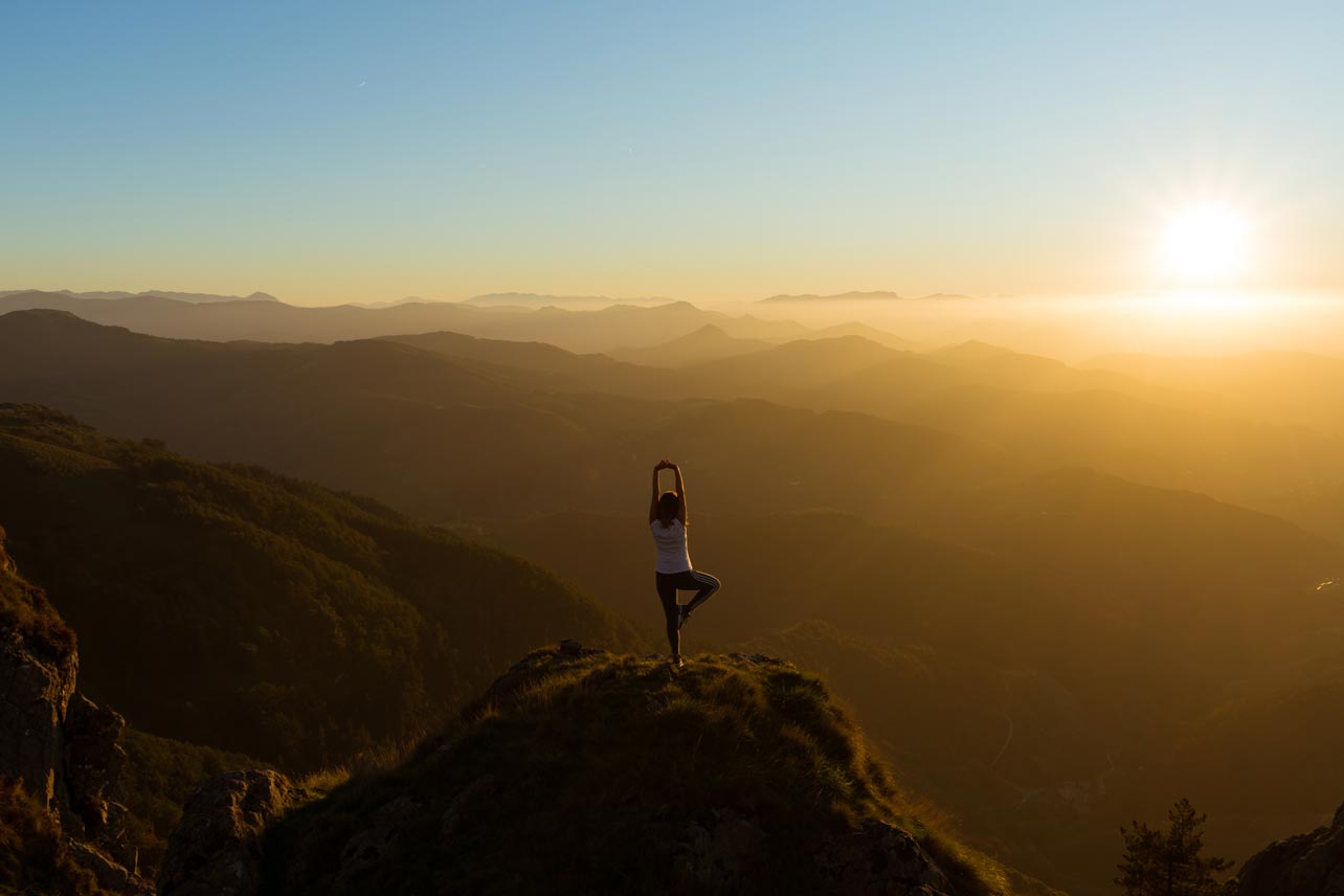 A woman in a yoga pose on a hilltop overlooking ridges of mountains at sunset